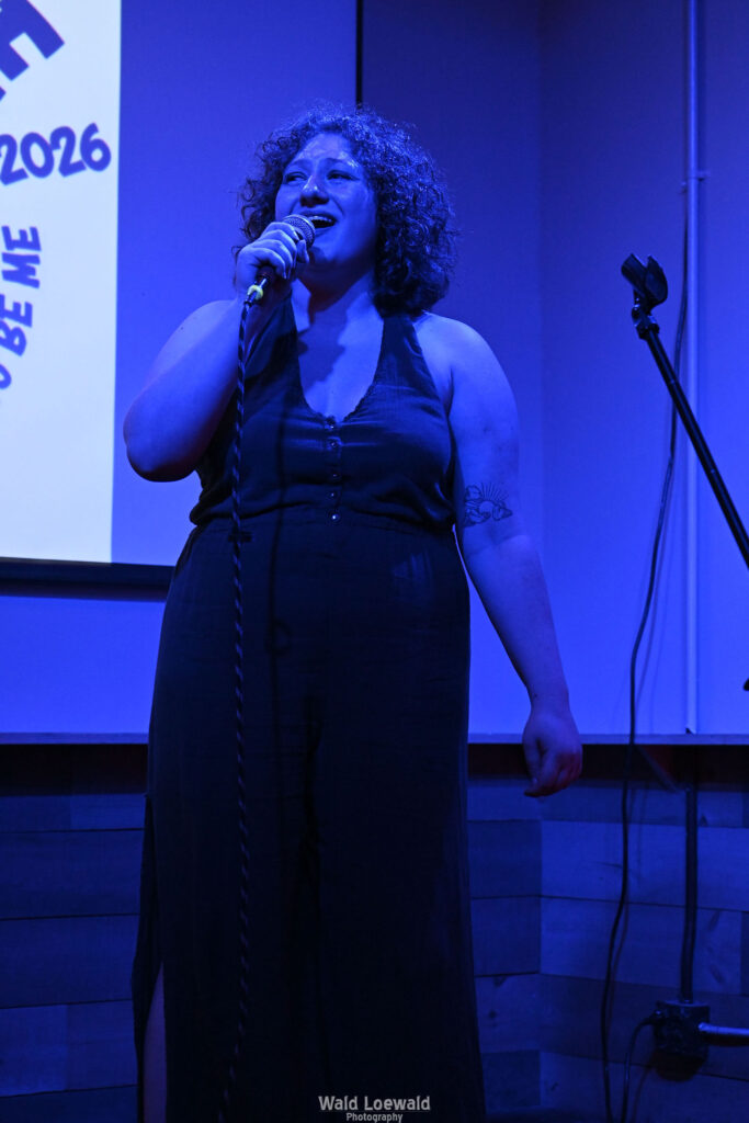 Performer in blue lighting holding a mic in front of Mad Pride 2026 logo
