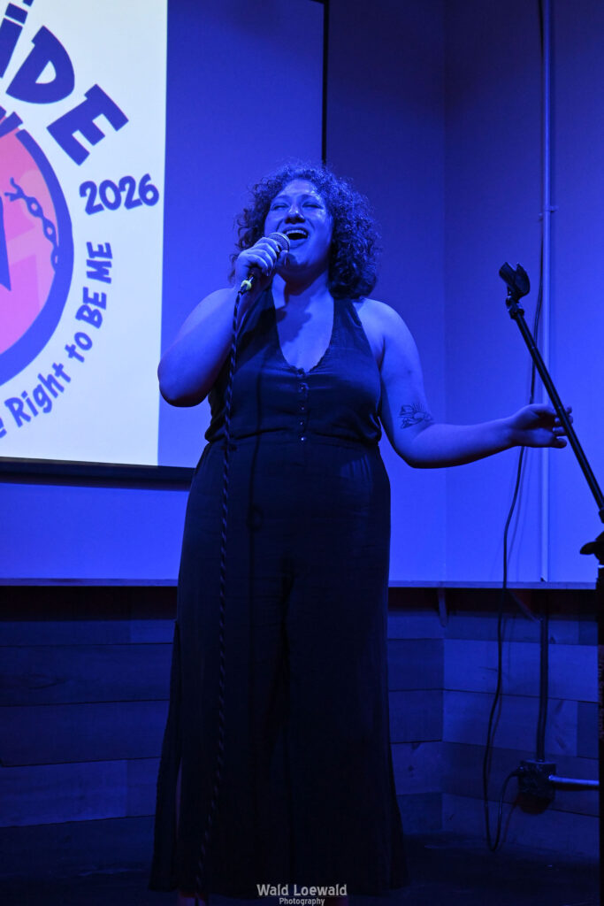 Performer in blue lighting holding a mic in front of Mad Pride 2026 logo