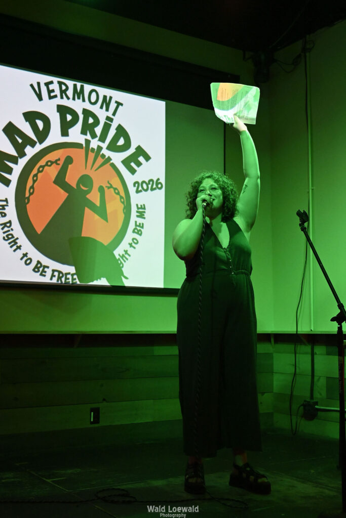 Performer in green lighting holding a mic in front of Mad Pride 2026 logo
