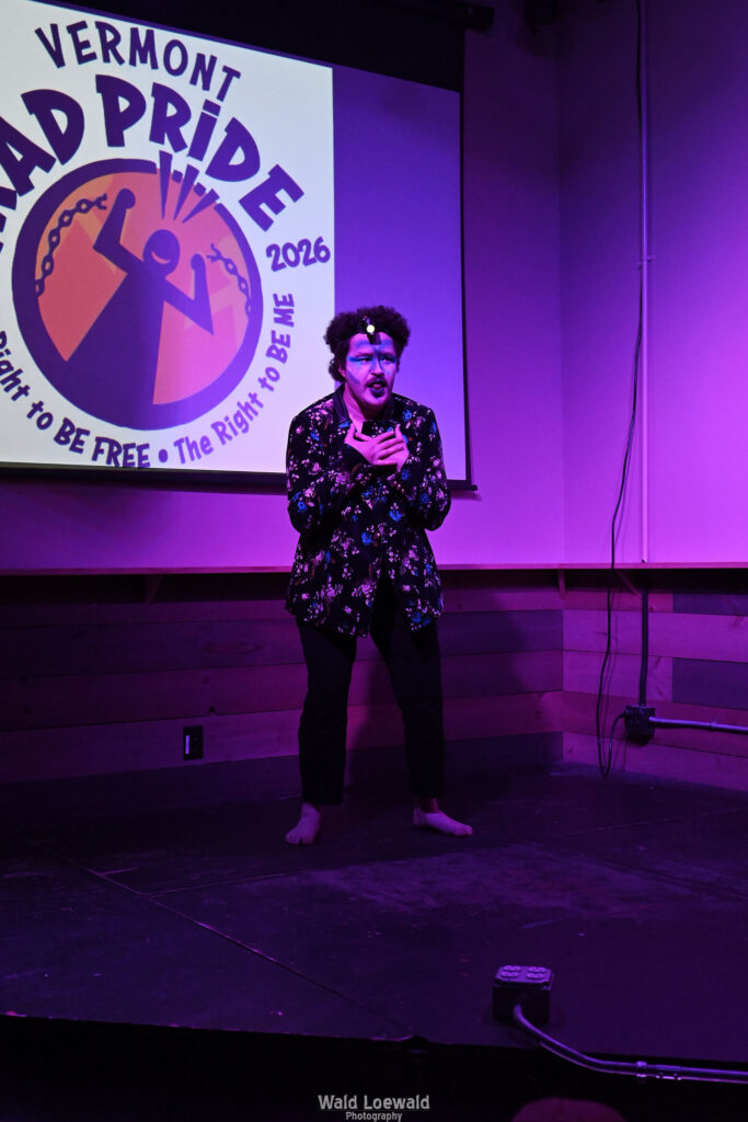 Performer on stage in costume in pink lighting in front of Mad Pride 2026 logo