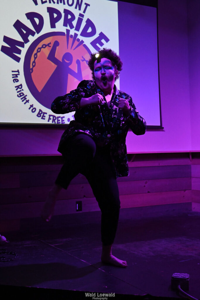 Performer on stage in costume in pink lighting in front of Mad Pride 2026 logo