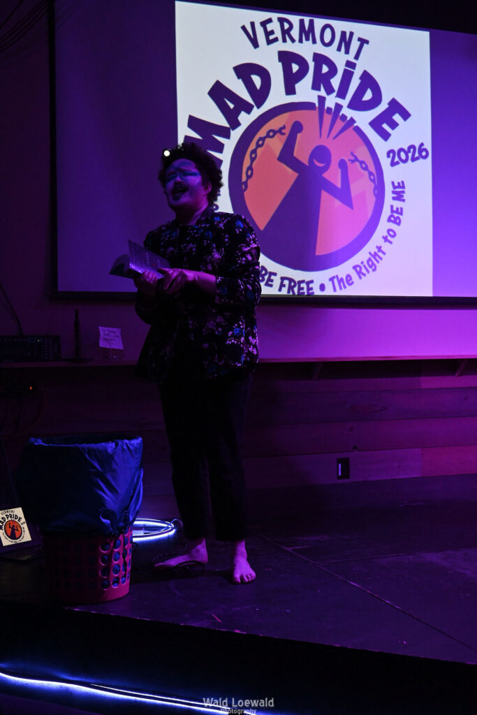 Performer on stage in costume in pink lighting in front of Mad Pride 2026 logo