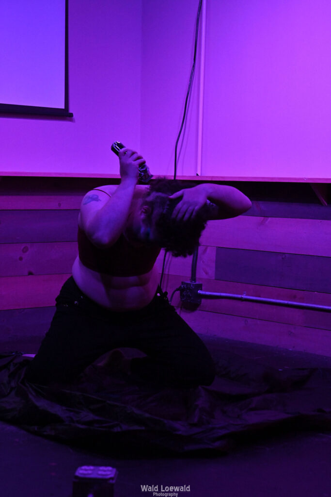 Performer shaving head on stage in pink lighting
